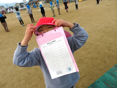 園庭で赤い帽子の園児が首から下げた記録カードを持ち上げて見せている写真