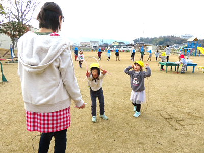園庭で職員と園児二人が一緒に縄跳びを跳んでいる様子の写真