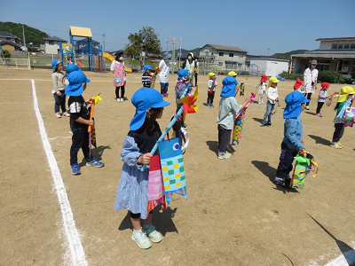 園児たちがカラフルなこいのぼりを片手に持ち園庭に整列している写真