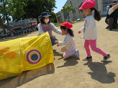 先生に付き添われながら黄色い鯉のぼりの形をしたトンネルをくぐる女の子2人の写真