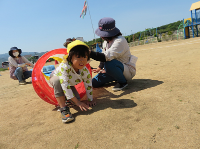 先生二人が鯉のぼりトンネルを支え、一人の男の子が勢いよく鯉のぼりトンネルから出てくる様子の写真