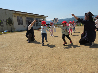 2人の先生が持った鈴を一生懸命ジャンプして鳴らそうとしている男の子と女の子の写真