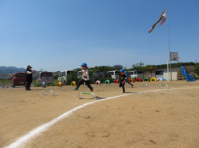 雲一つない青空のもと、園庭に設置されたハードルを元気に飛び越えている園児たちの写真