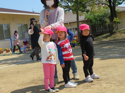 マイクを持った先生と経っている3人の園児たちの写真