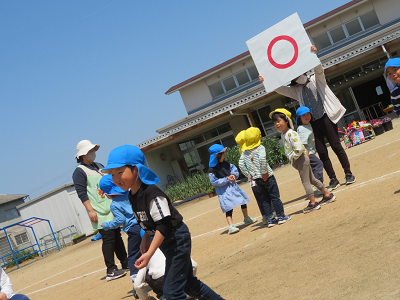 大きな「丸」が書かれたプレートを持った先生のところへ集まる園児たちの写真