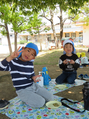 木陰でレジャーシートを敷き、かしわもちを食べる2人の園児の写真