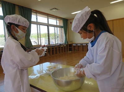 給食用の白衣を着た女の子二人がボウルに入ったホットケーキの生地をこねている様子の写真