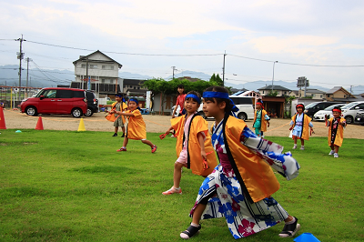 色鮮やかな緑の芝生の上で、黄色い法被を着た園児たちが両手を広げ、身体全体を使って元気にソーラン節を踊っている写真