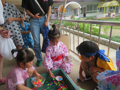 ピンクの浴衣を着た女の子2人が金魚すくいのポイを片手に、真剣な表情でスーパーボールすくいをしている写真