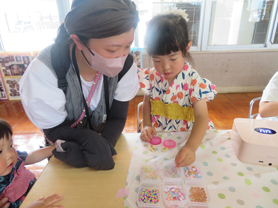 一人の女性と一緒に浴衣姿の女の子が色んな種類のビーズが入った箱からビースを選びキーホルダーづくりをしている写真