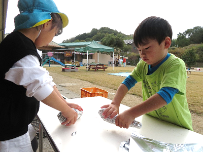 外に設置された机の上で女の子と男の子がさつまいもをアルミホイルで包んでいる写真