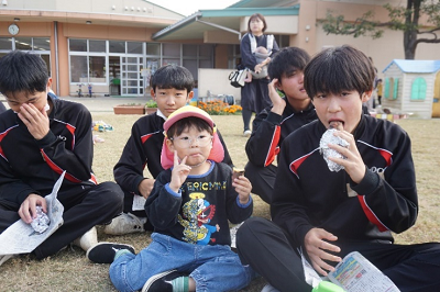4人の男子中学生と一緒に座って焼き芋を美味しそうに食べながらピースサインをする男の子の写真