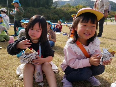 芝生の広場に座り込んで熱々の焼き芋を美味しそうに食べている2人の女の子の写真