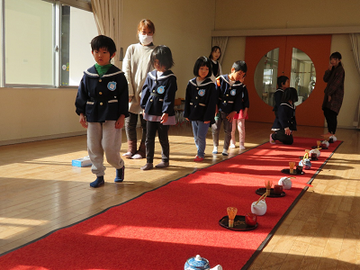 赤い敷物の上に並べた道具の横を園児たちが歩いている様子の写真