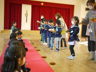園児たちが赤いカーペットに正座で座っている園児たちに順番にお茶を運んでいる様子の写真