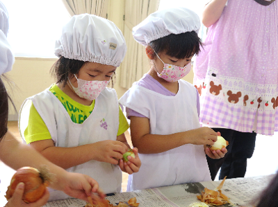 調理帽をかぶった子どもたちが玉ねぎや野菜の皮を上手にはいでいる写真