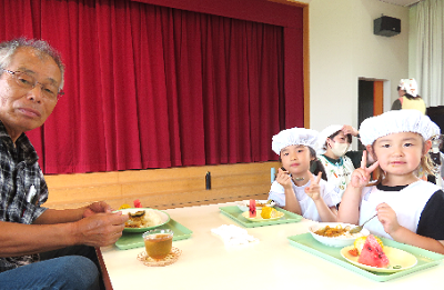 赤い幕の前で地域の方と園児たちがカレーを前に並んで座り、ピースをしている写真