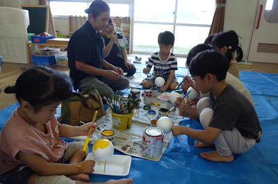 保育室のビニールシートの上で園児たちが丸い土台に絵の具を塗っている写真