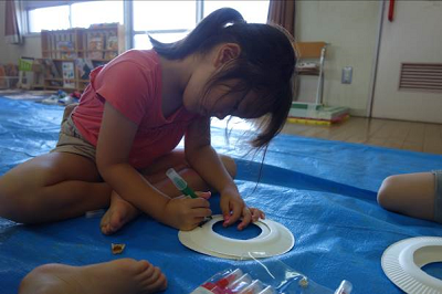 保育室の床に座った園児が紙皿にペンで模様を描いている写真