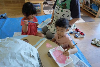保育室で幼い園児たちが大きな段ボールの土台にピンクの絵の具を塗っている写真