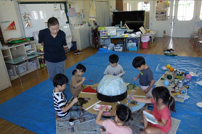 保育室の中央で教師と園児たちが大きな立体作品の周りに集まり色を塗っている写真