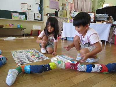 保育室の床で園児たちが色とりどりの靴下をつなげた長い作品に飾り付けをしている写真