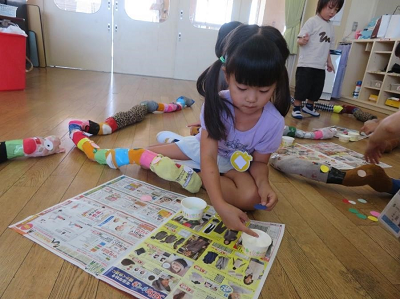 保育室で園児が広告紙の上で丸いパーツを用意して靴下のヘビの飾りを作っている写真