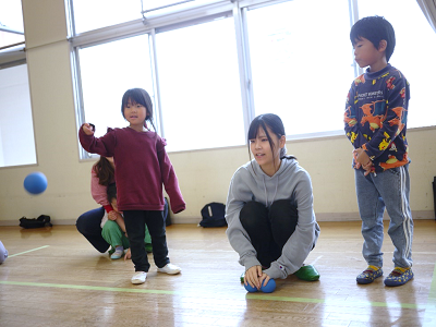 体育館で女の子が青いボールを投げ先生や友達が見守っている写真