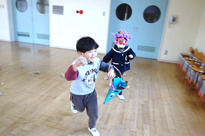 教室で園児が鬼のお面をつけた友達を追いかけながら豆まきをしている写真