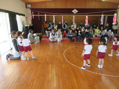 遊戯室で見守っている保護者の前で集まっている園児たちの写真