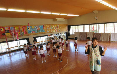 保育士が前で見本を見せ、それに合わせて園児たちが遊戯室で踊っている様子の写真