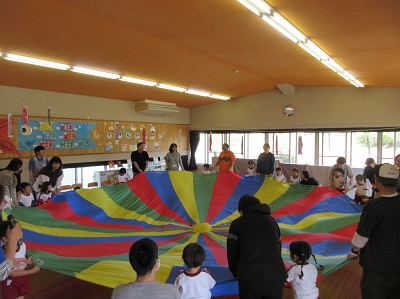 園児と保護者が遊戯室で大きなカラフルなバルーンを広げている写真