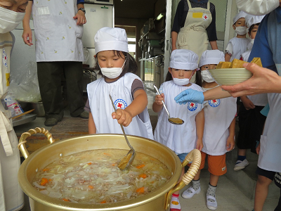 園児が沢山の具材が入った大きな鍋にお玉でそっとカレールーを入れている様子の写真