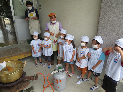 園児たちが屋外で調理中のカレーの入った大鍋の前に並んでいる写真