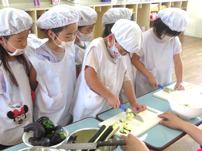調理室でエプロンと帽子をつけた子どもたちが野菜を包丁で切っている写真