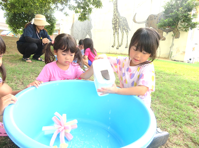 大きなたらいに液体を注いで水遊びをしている女の子とそれを見守る友だちの写真