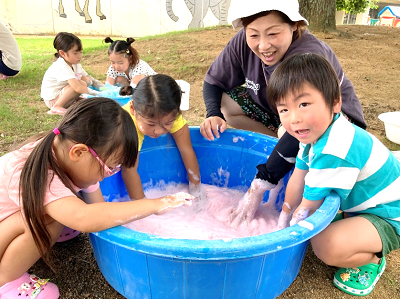 ピンク色の泡が入ったたらいで遊ぶ子どもたちとそばで見守る保育者の写真