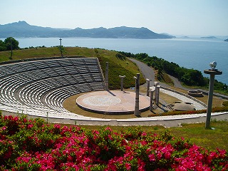 手前に赤い花と奥に海が見える場所に扇状の観客席の中央にステージがある写真