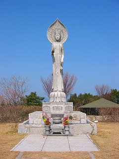 茶色い芝生の上に立てられた縦に細長い灰色の観音像の写真