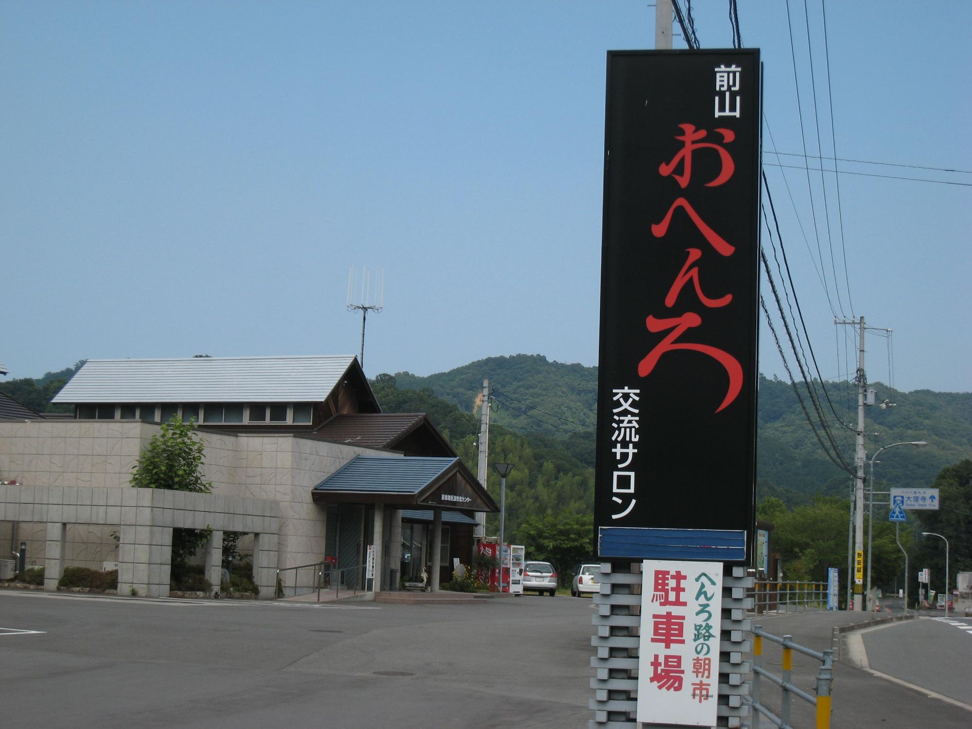 駐車場の横に「おへんろ」と書かれた黒い縦長の看板が立ち、その奥に建物と山並みが見える写真