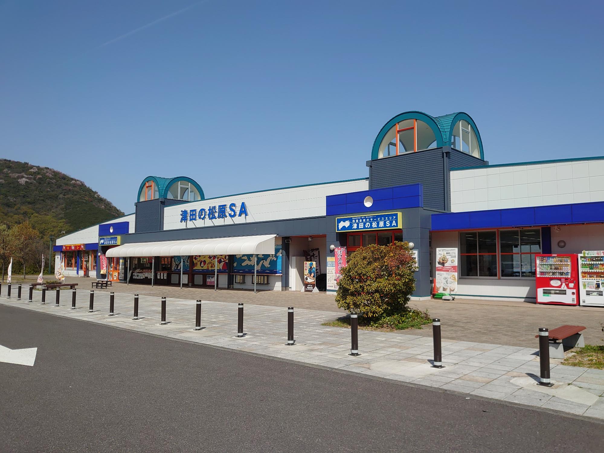 青空の下、駐車場に面して青と白の外観のサービスエリアが建っている写真