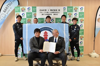 大きなSONIOのフラッグを持った5人の前に調印書を掲げた2人が座っている写真