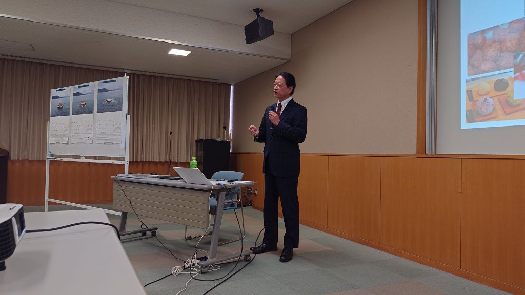 会議室の前方でスーツ姿の男性講師がスクリーンを前に説明している写真