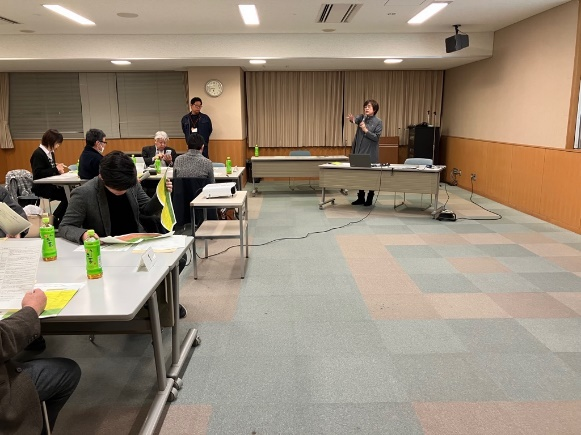 会議室で話者の女性が立って説明し、参加者が資料を見ながら話を聞いている写真