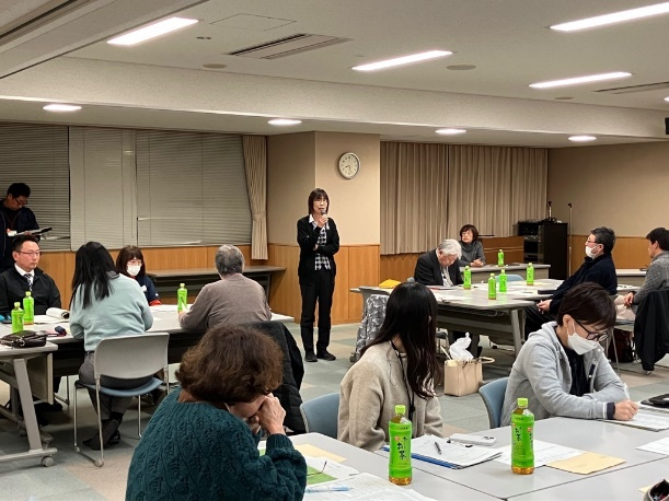 会議室の中央で立って話す女性の説明を、参加者たちが聞いている写真