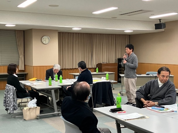 会議室の一角で男性がマイクを持って説明し、参加者が資料を確認している写真