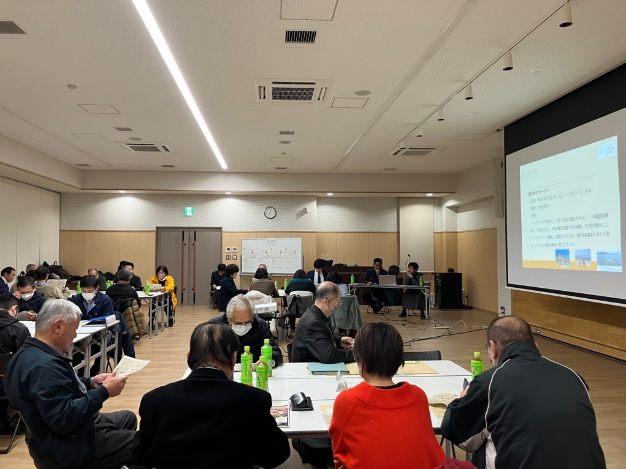 会議室でスクリーンの説明を聞きながら資料を見る参加者たちの写真