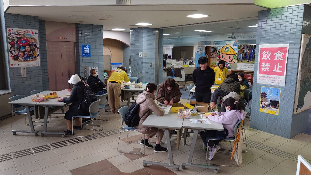 館内の休憩スペースのテーブルで親子連れや来場者たちがスタッフに教わりながら工作体験をしている写真