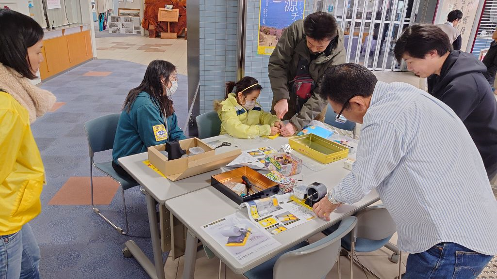 館内のテーブルで職員と保護者に囲まれた子どもが小さな部品を手作業で組み立てている様子を間近から写した写真