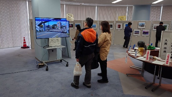 展示室の一角で来場者たちがモニターに映るまちの映像や周囲の作品パネルを眺めている写真